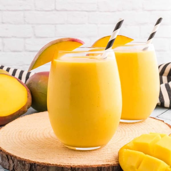 Two glasses of mango lassi with mango slices and striped straws on a wooden board.