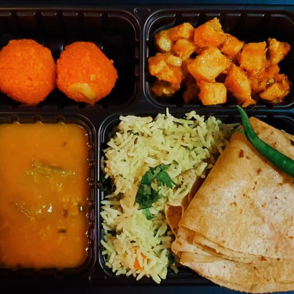 Indian thali with dal, rice, roti, paneer, and two orange desserts in a black takeout container.