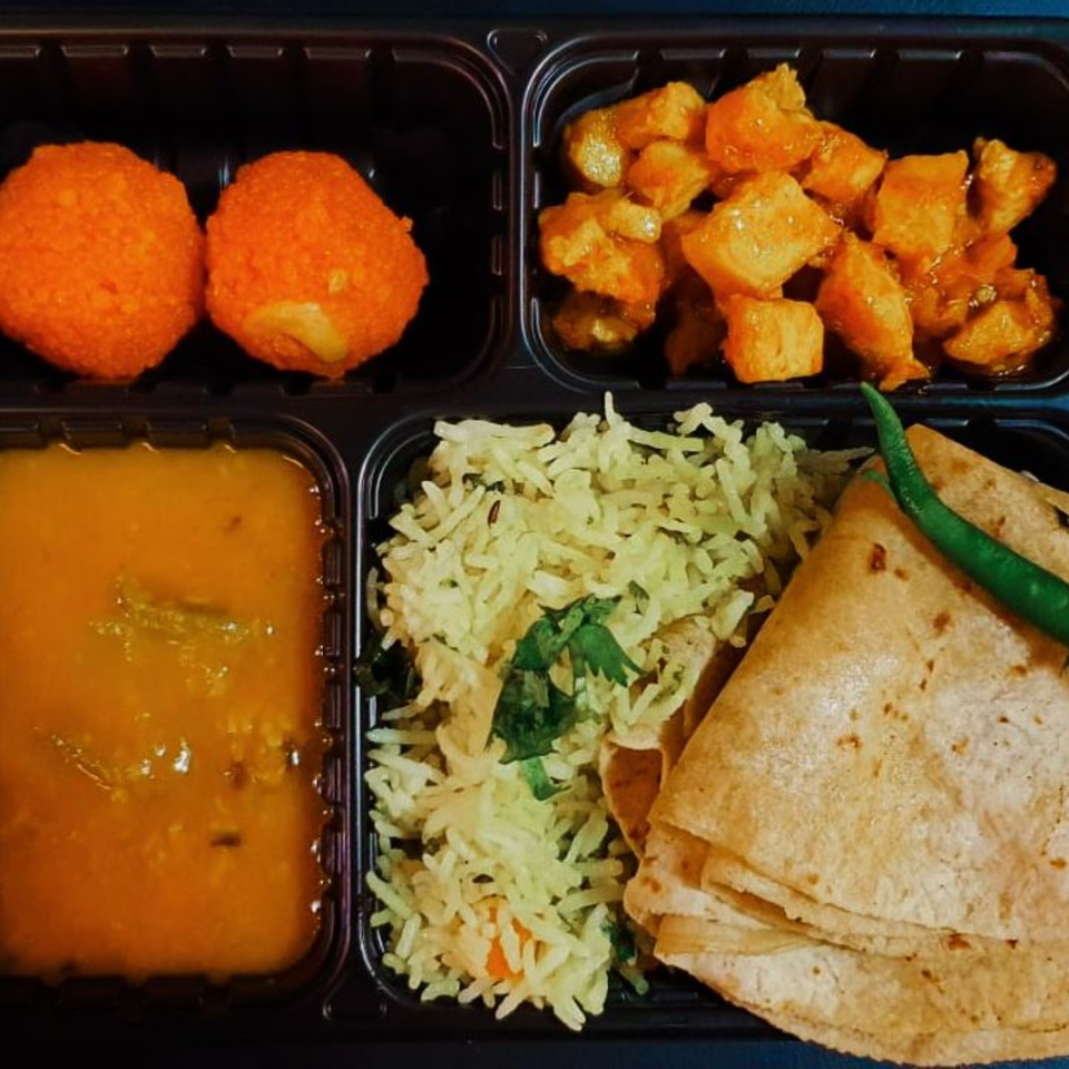 Indian thali with dal, rice, roti, paneer, and two orange desserts in a black takeout container.
