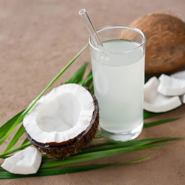 Fresh coconut water in a glass with a straw, surrounded by whole and cracked coconuts and green leaves.