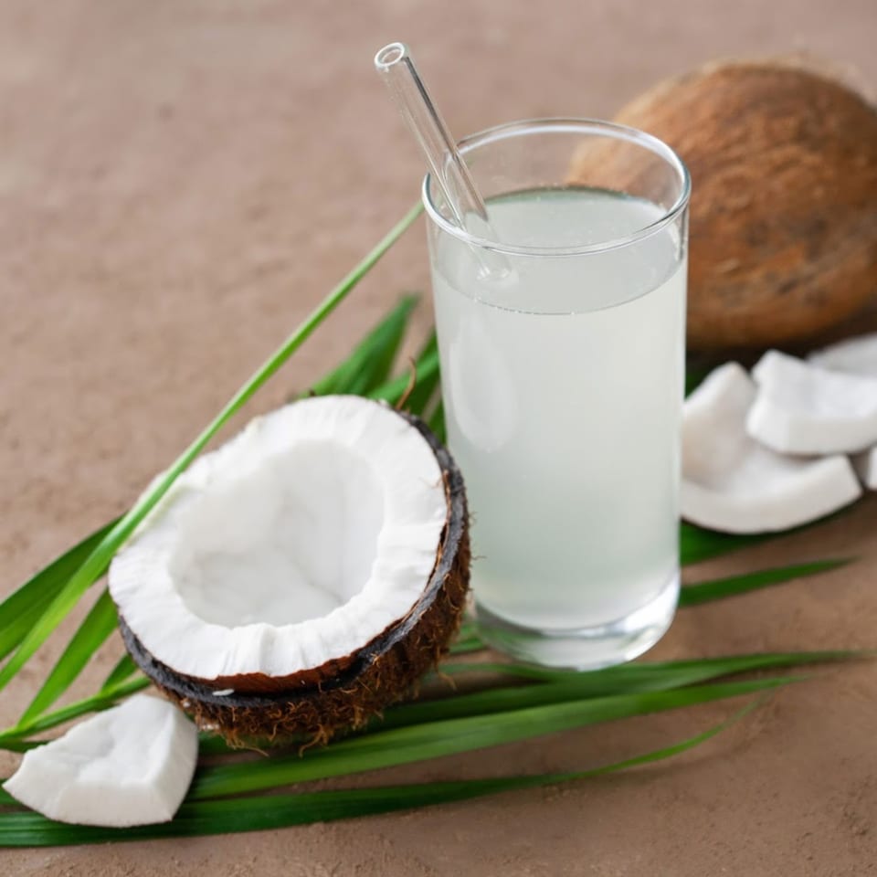 Fresh coconut water in a glass with a straw, surrounded by whole and cracked coconuts and green leaves.