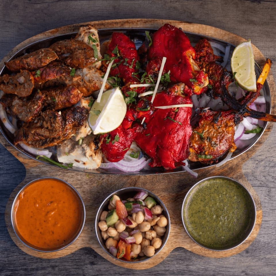 A large platter of various Indian grilled meats, including red tandoori chicken, with three dipping sauces.