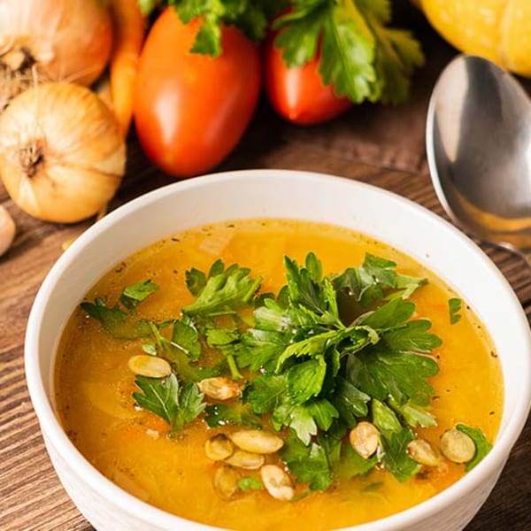 A bowl of golden Indian soup, garnished with fresh parsley and pumpkin seeds, surrounded by vegetables.
