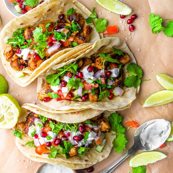 Three Indian chicken tacos with pomegranate, cilantro, and white sauce on a brown paper background.